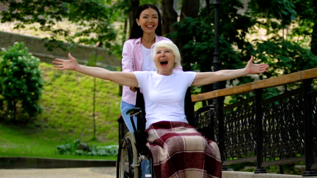 Lächelnde-Freiwillige-im-Park-mit-inspirierten-alten-Frau-im-Rollstuhl-heben-Hände