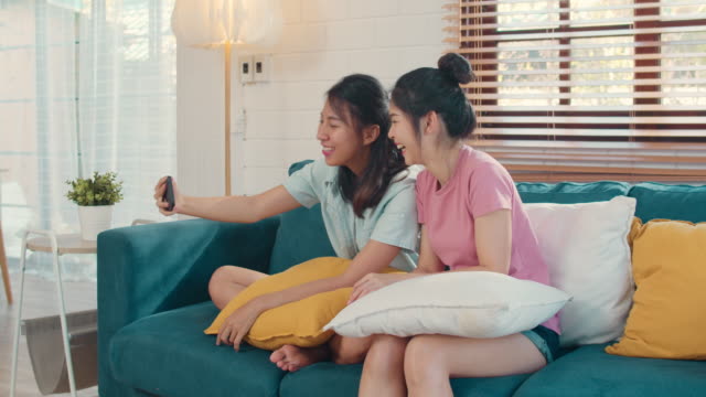 Lesbian-influencer-couple-using-mobile-phone-record-vlog-video-upload-on-social-media-while-lying-sofa-in-living-room.