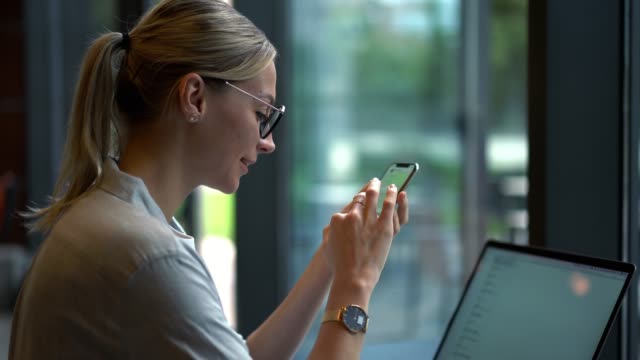 Successful-female-entrepreneur-enjoying-chatting-via-smartphone-app-while-sitting-at-desk-with-computer