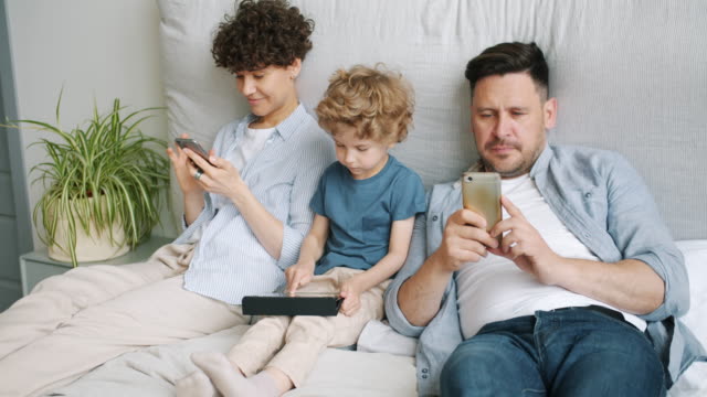 Mann-und-Frau-mit-Smartphones,-während-Kind-spielen-Spiel-auf-Tablet-im-Bett-zu-Hause