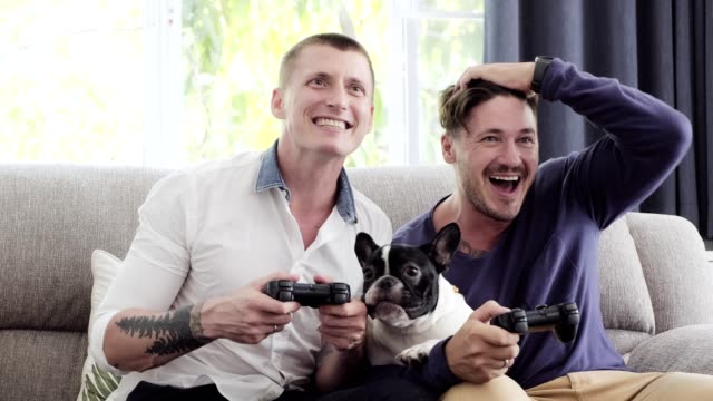 Gay-couple-relaxing-on-couch-with-dog-playing-games.-Loses.