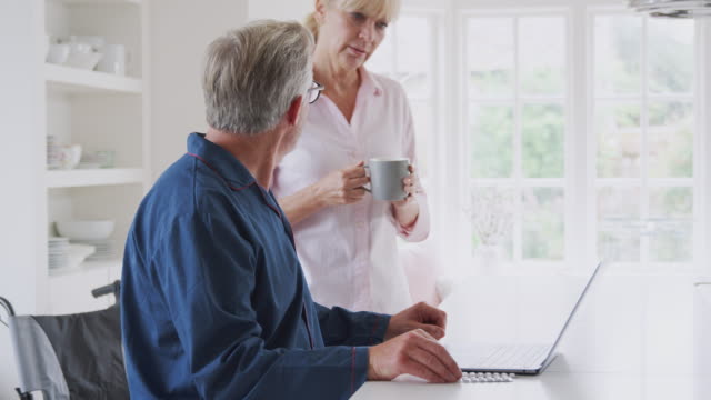 Senior-Couple-With-Man-In-Wheelchair-Looking-Up-Information-About-Medication-Online-Using-Laptop