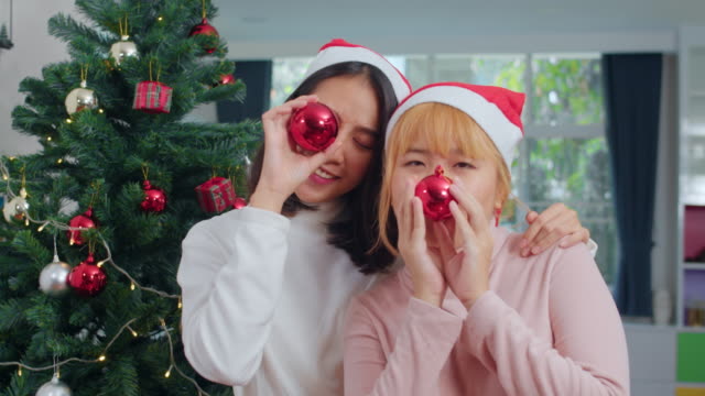 Pareja-lesbiana-asiática-celebran-festival-de-Navidad.-LGBTQ-adolescente-mujer-usan-sombrero-de-Navidad-relajarse-feliz-jugar-bola-de-Navidad-disfrutar-de-las-vacaciones-de-invierno-de-Navidad-juntos-en-la-sala-de-estar-en-casa.-Disparo-a-cámara-lenta.