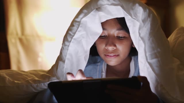 Asiática-niña-usando-tableta-bajo-manta-en-el-dormitorio-por-la-noche