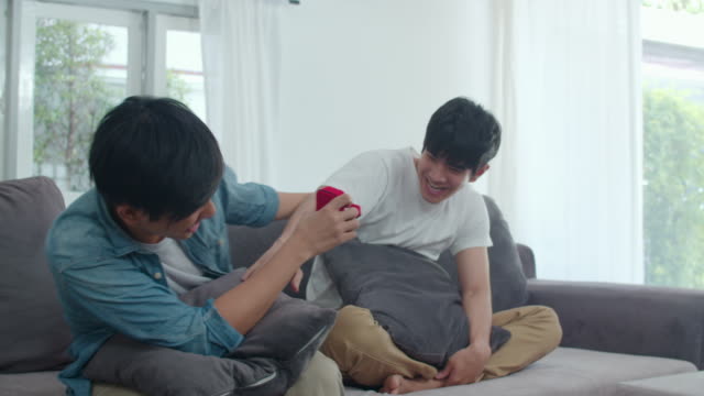 Junge-asiatische-homosexuell-Paar-schlagen-zu-Hause,-Teen-korean-LGBTQ-Männer-glücklich-lächeln-haben-romantische-Zeit,-während-Vorschlag-und-Ehe-Überraschung-tragen-Ehering-im-Wohnzimmer-im-Haus-Konzept.