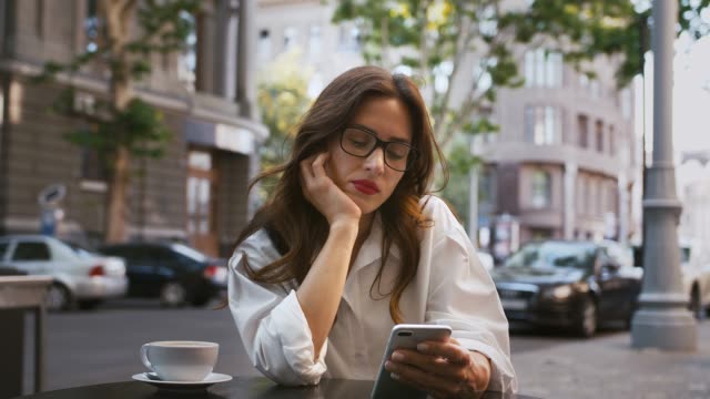 Business-lady-in-glasses,-white-shirt.-Sitting-at-table-with-cup-of-coffee-in-roadside-cafe.-Browsing-news-on-smartphone,-working-online.-Close-up