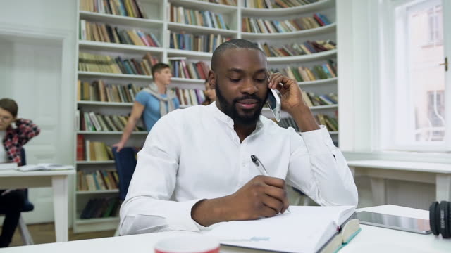 Zeitlupe-von-sympathischen-zufriedenen-bärtigen-afrikanischen-amerikanischen-Kerl,-der-Notizen-in-Copybook-während-seiner-Telefon-Gespräch-in-der-Bibliothek