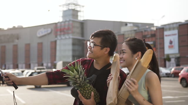 Divertido-joven-par-vietnamita-de-hombre-y-mujer-haciendo-selfie-con-peanapple-y-pan-largo-cerca-del-supermercado