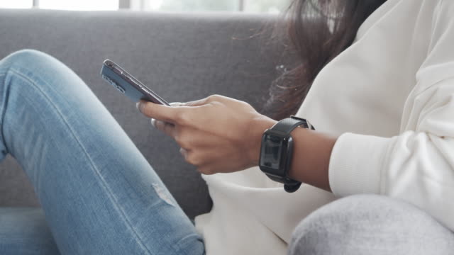 Close-up-hands-asian-female-using-smartphone-checking-social-media-and-shopping-online-store-while-laying-on-a-sofa-in-the-living-room-at-home-during-pandemic-coronavirus.