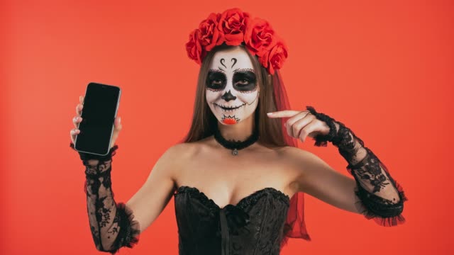 Día-de-Muertos,-mujer-con-maquillaje-cráneo-sosteniendo-el-teléfono-inteligente,-señalando-a-su-pantalla-por-el-dedo-índice-y-sonriendo-posando-sobre-fondo-rojo,-Halloween