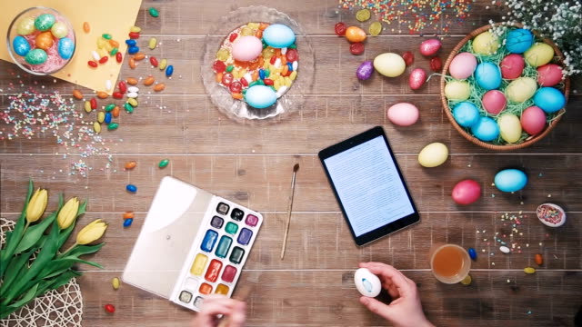 Huevo-de-Pascua-de-hombre-pintura-y-tableta-digital-se-encuentra-en-mesa-decorada-con-vista-superior-de-huevos-de-Pascua