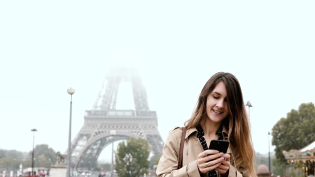 Young-beautiful-woman-standing-near-the-Eiffel-tower-in-Paris,-France-and-using-the-smartphone-in-foggy-morning