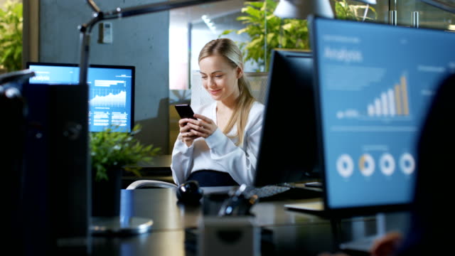 In-the-Office-Beautiful-Businesswoman-Uses-Her-Mobile-Phone-and-Smiles-Attractively.-She-Sits-at-Her-Desk.