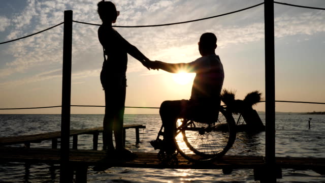 silhouette-of-Lovers-on-wheelchair-standing-to-quay-on-background-of-water-and-sky-in-sunset