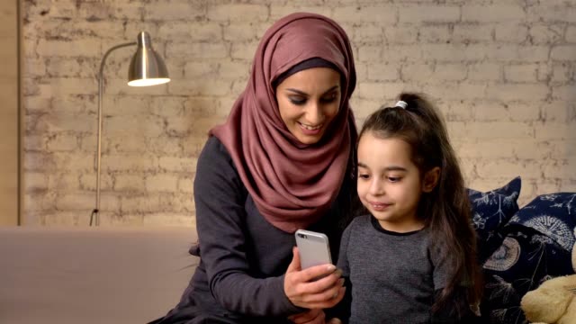 Joven-madre-hermosa-en-hijab-con-niña-en-el-sofá,-sonrientes,-aplicaciones-smartphone,-hace-selfie-caricias,-pequeña-niña-con-oso-de-peluche,-confort-hogar-concepto-de-familia-feliz-en-el-fondo-50-fps