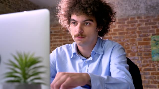 Serious-nerdy-businessman-with-curly-hair-typing-on-laptop-and-sitting-in-modern-office,-programmer-concept