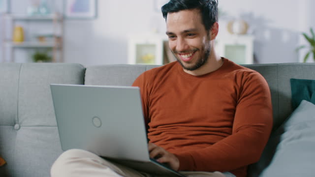 Fröhlicher-junger-Mann-sitzt-auf-einem-Sofa-hält-Laptop-auf-dem-Schoß,-browst-durch-das-Internet,-soziale-Netzwerke,-macht-e-Shopping.-Mann-zu-Hause-mittels-Laptop-beim-Sitzen-auf-einer-Couch.
