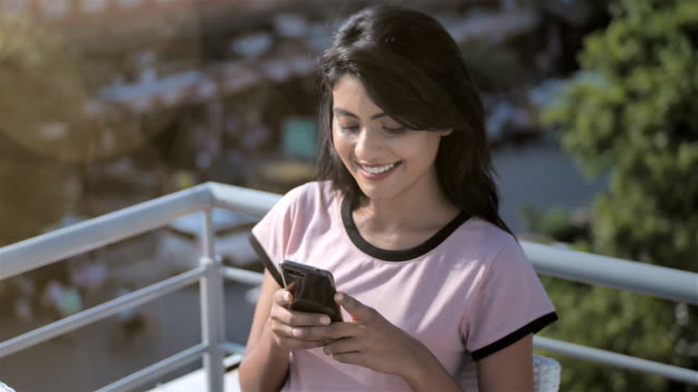Eine-schöne-junge-Frau-lächelnd,-während-Sie-mit-dem-Handy-oder-Smartphone-sitzt-in-einem-Café-auf-der-Dachterrasse-vor-der-belebten-Straße.