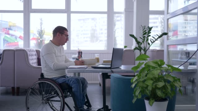 e-learning,-senior-male-disabled-in-wheelchair-wearing-glasses-reads-book-and-uses-laptop-sitting-at-a-table-in-a-cafe