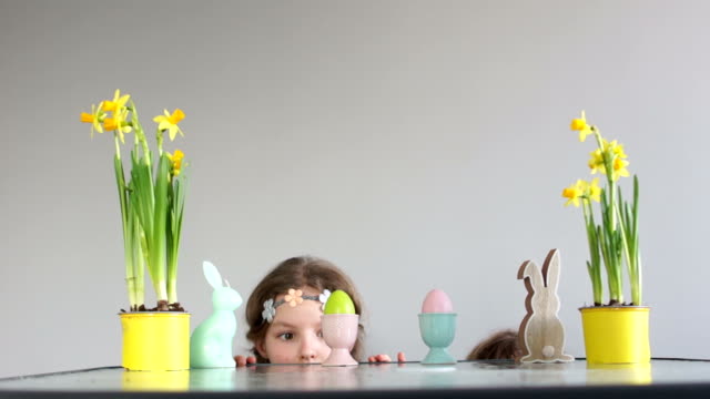 Funny-children,-a-boy-and-a-girl-peeking-from-the-table-with-Easter-decor.-Search-for-Easter-eggs