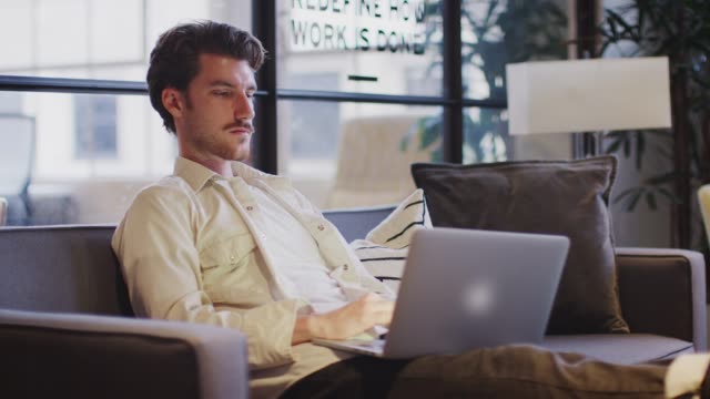 Young-man-working-on-a-laptop-sitting-with-his-feet-up-in-an-office-lounge-area,-side-view