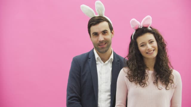 Young-beautiful-couple-standing-on-a-pink-background.-With-hackneyed-ears-on-the-head.-During-this-man-and-his-wife-are-looking-into-the-camera.