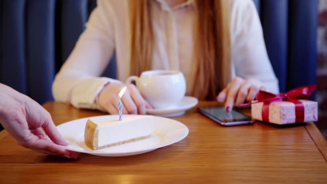 Close-up-of-waitress-serving-cake-with-burning-candle-for-woman-having-tea-and-browsing-smartphone-alone-in-cafe.-Birthday-gift-box-placed-on-table