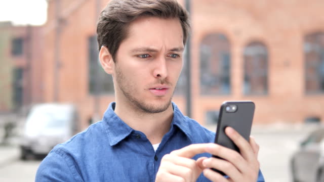 Outdoor-Portrait-of-Young-Man-Upset-by-Loss-while-Using-Smartphone