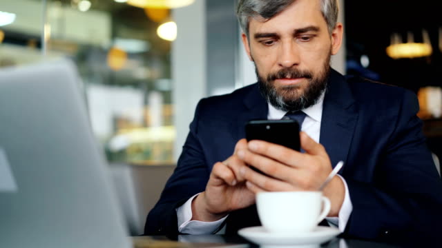 Bearded-office-worker-in-suit-using-smartphone-during-coffee-break-in-cafe