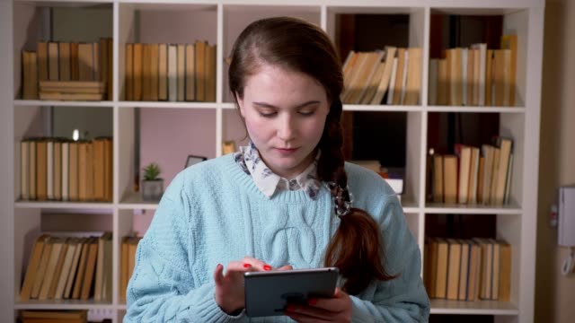 Nahaufnahme-Porträt-von-jungen-hübschen-Studentin-SMS-auf-dem-Tablet-Blick-auf-die-Kamera-in-der-Universitätsbibliothek-in-Innenräumen