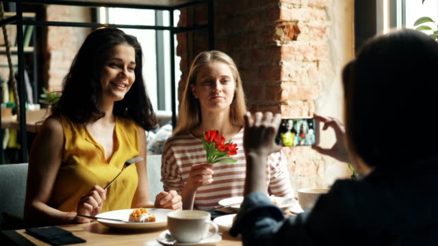 Mujeres-despreocupadas-posando-para-la-cámara-del-teléfono-inteligente-en-el-café-sosteniendo-la-flor-divirtiéndose