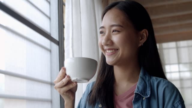 Cierre-atractivo-retrato-sonriente-joven-asiática-mujer-sosteniendo-taza-de-café-de-pie-junto-a-la-ventana-en-la-oficina-en-casa.