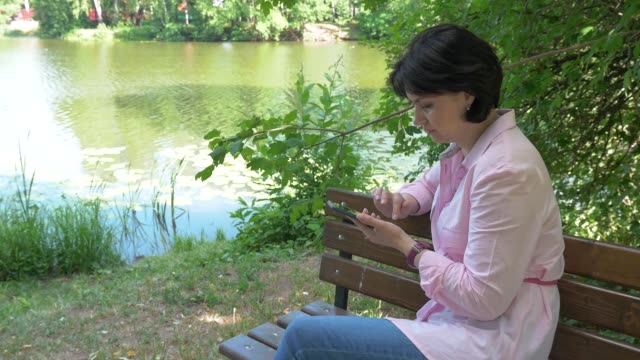 Eine-brünette-Frau-in-einem-Park-auf-einer-Bank-schaut-auf-ein-Foto-auf-ihrem-Smartphone
