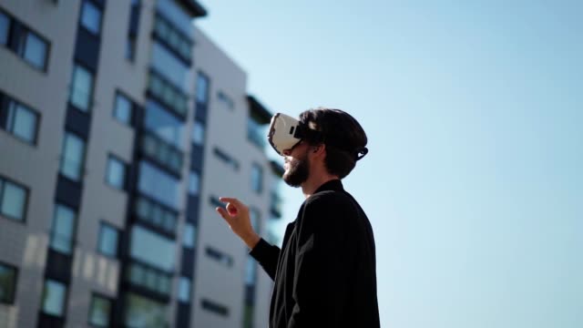 Panning-slow-motion-shot-of-young-man-wearing-vr-headset-standing-outdoors-in-city-street-and-exploring-data-in-sunlight