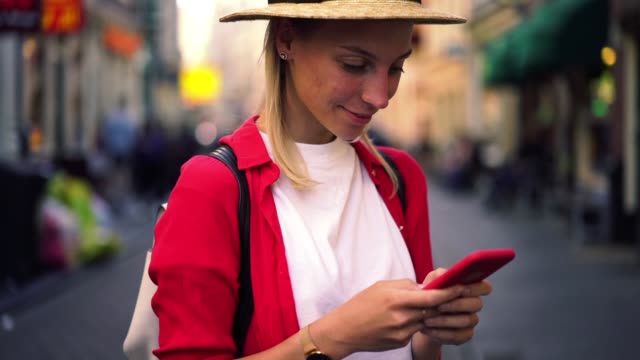 Slow-motion-effect-of-caucasian-female-traveler-in-hat-looking-at-camera-while-holding-mobile-phone-on-street