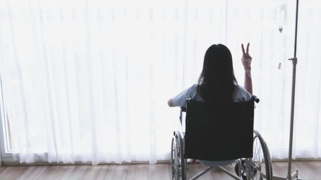 Asiatische-Frauen-Patient,-behinderte-Bein-sitzen-im-Rollstuhl-und-zeigt-Ermutigung-Symbol-mit-zwei-Fingern.