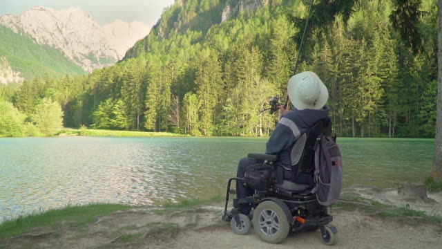 Cámara-lenta-de-pescador-discapacitado-en-una-silla-de-ruedas-eléctrica-pescando-en-un-hermoso-lago-cerca-del-bosque-y-la-montaña-en-la-parte-posterior