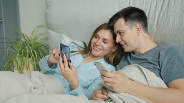 Atractivo-hombre-y-mujer-usando-teléfonos-inteligentes-en-la-cama-hablando-sonriendo-en-casa