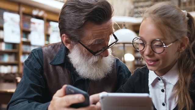 Intelligenter-Senior-Mit-zerknitterter-Gesicht-und-grauem-Bart-zeigt-etwas-auf-seinem-Handy-zu-seiner-lächelnden-Teenager-Enkelin,-während-sie-in-der-Bibliothek-sitzen
