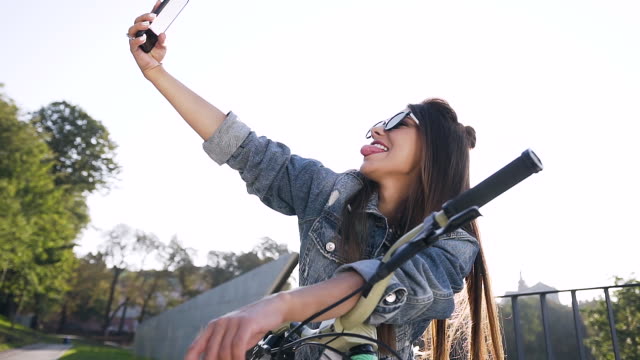 Wunderschöne-fröhliche-Mädchen-in-modischen-Sonnenbrille-und-stilvolle-Kleidung-sitzen-auf-dem-Fahrrad-und-machen-lustige-Selfie-unter-Sonnenlicht