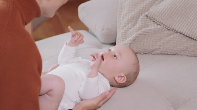 Close-up-of-the-baby-lying-on-its-back-on-the-couch,-smiling-and-happy,-shaking-hands,-looking-at-its-mother,-who-kneels-by-the-baby,-talks-to-it-and-smiles