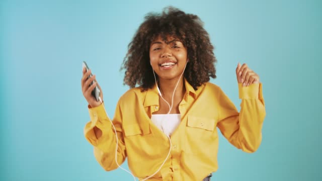 Dark-skinned-woman-listening-to-music-through-earphones-connected-to-smartphone,-dancing-and-smiling-posing-on-blue-background