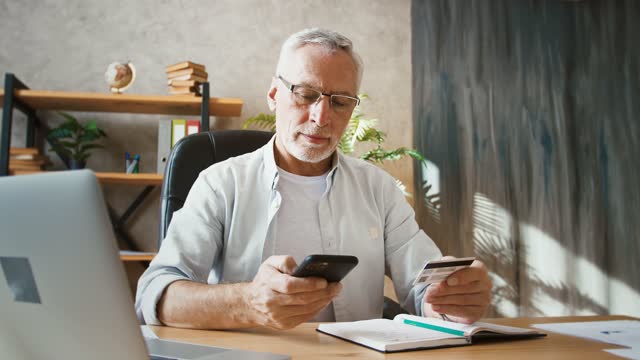 Hombre-de-la-tercera-edad-sosteniendo-tarjeta-de-crédito-e-ingresando-su-número-en-el-teléfono-móvil,-comprando-en-línea-trabajando-en-la-oficina-en-casa