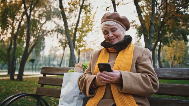 Rentnerin-schaut-auf-Bildschirm-ihres-Smartphones-und-lächelt,-während-sie-auf-der-Bank-im-Herbstpark-sitzt