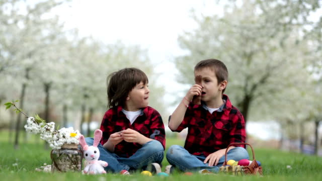 Two-children,-boy-brothers,-eating-chocolate-bunnies-and-having-fun-with-easter-eggs-in-the-park,-beautiful-spring-blooming-garden