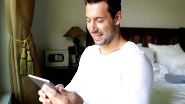 Young-Man-Using-Tablet-Computer-Drink-Coffee-Smiling-Guy-Chatting-Online-In-Bedroom