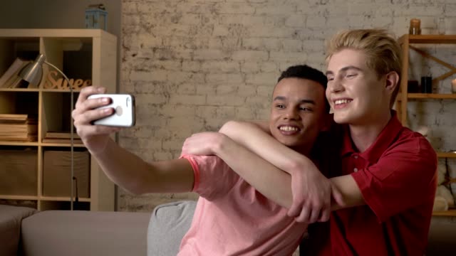 Two-international-gay-friends,-sitting-on-the-couch,-hugging,-making-selfies,-laughing.-Homeliness,-romantic-evening,-background,-hugs,-happy-LGBT-family-concept.-60-fps