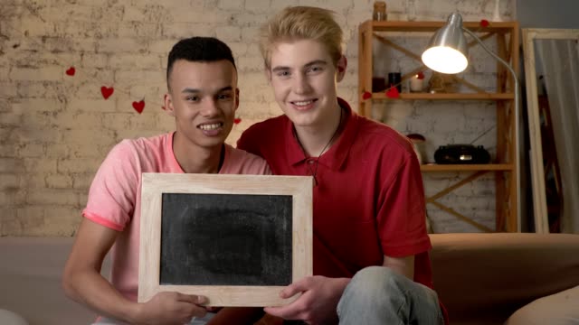 A-happy-international-gay-couple-is-sitting-on-the-couch-and-holding-a-empty-sign.-Home-comfort-on-the-background.-60-fps