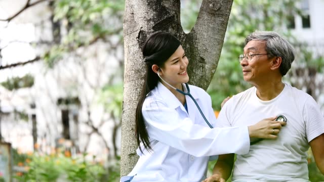 Young-female-doctor-using-the-stethoscope-listen-to-elderly-patient-heartbeat-in-hospital-garden
