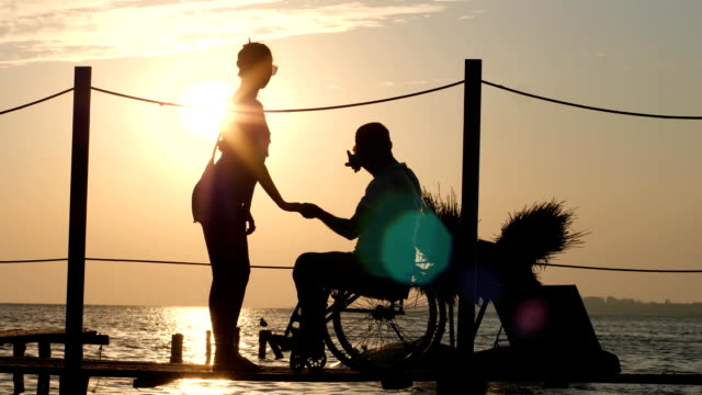 Silhouette-der-behinderte-Mensch-mit-Geliebte-Frau-auf-See-Pier-in-Abend-Abendrot-auf-Hintergrund-der-orangefarbenen-Himmel-und-Wasser